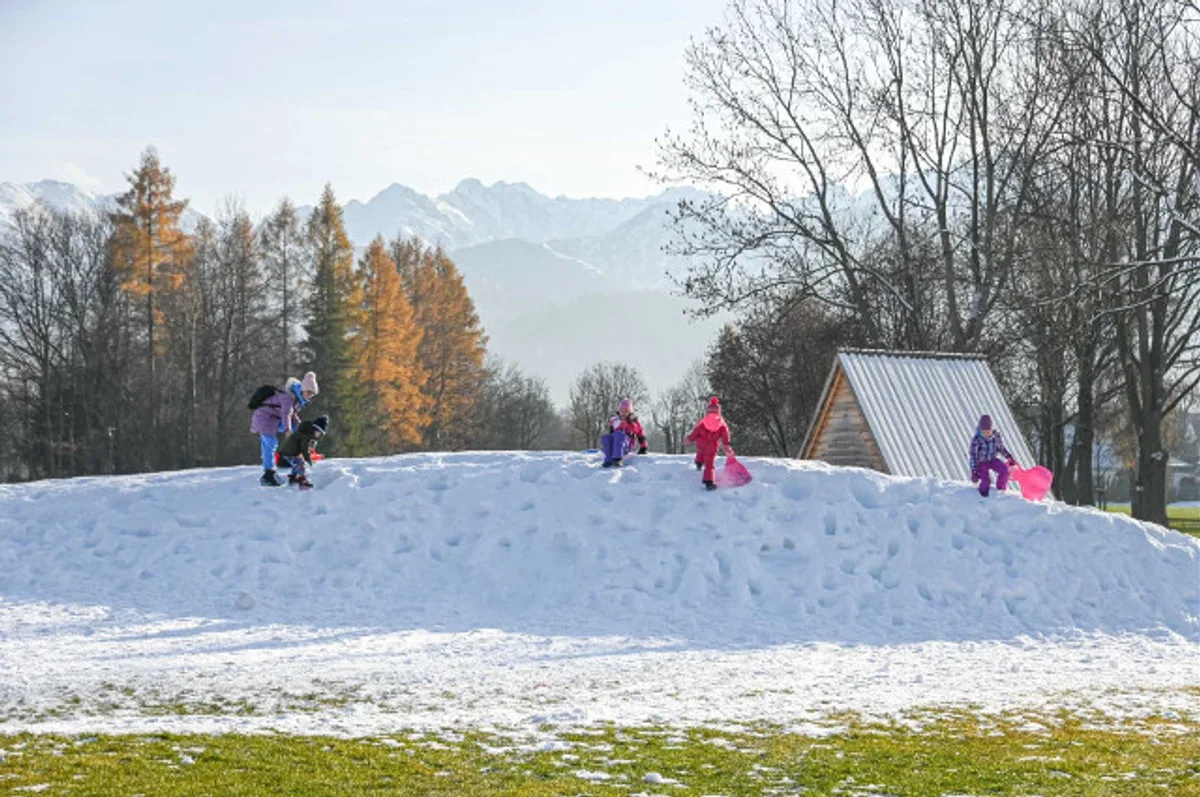 Dzieci z sankami na usypanej śnieżnej górce na Równi Krupowej w Zakopanem, w tle ośnieżone Tatry